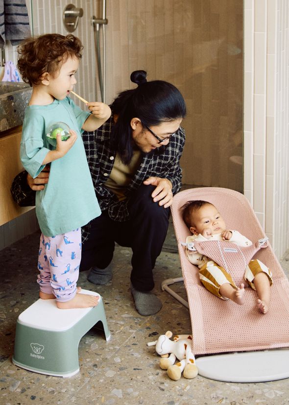 An adult kneels beside a baby in a pink bouncer. A toddler stands on a stool, holding a toy, in a bathroom setting.