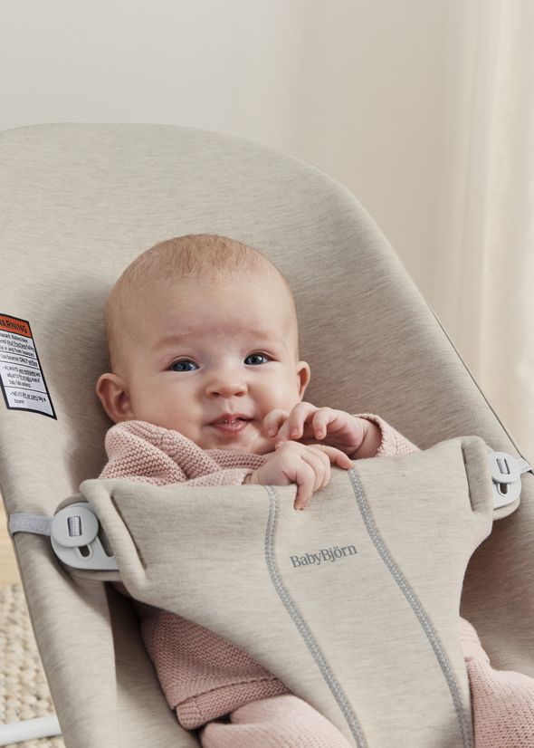 A baby in a pink outfit sits in a beige BabyBjörn bouncer, looking curiously forward with hands near the mouth.