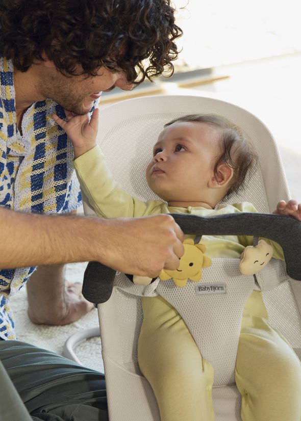 A baby in a yellow outfit sits in a bouncer, reaching up to touch a person's face. The person leans in, smiling, with a plaid scarf around their neck.