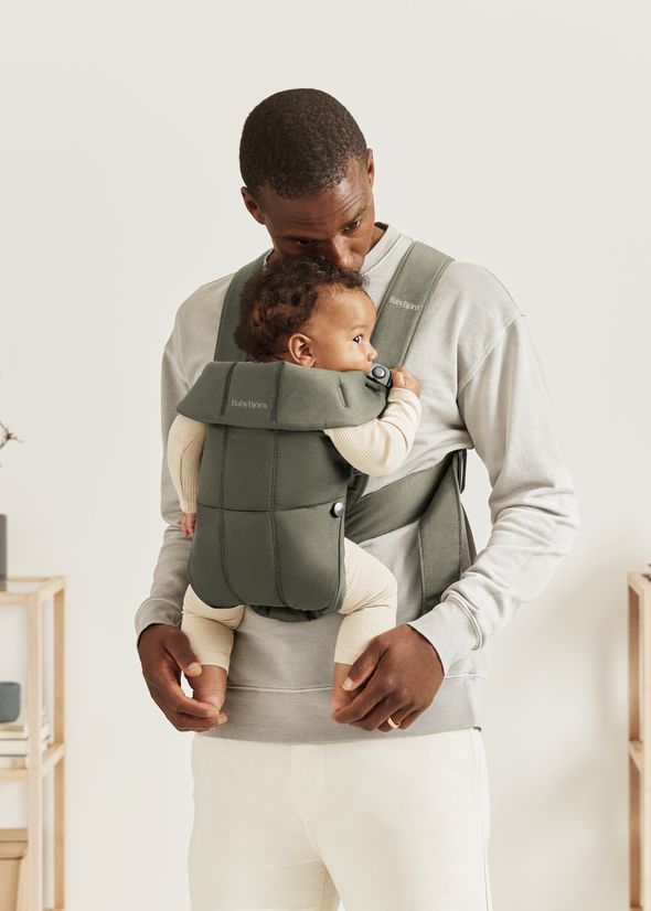 A man stands holding a baby in a green baby carrier, both dressed casually; the man gazes affectionately at the baby.