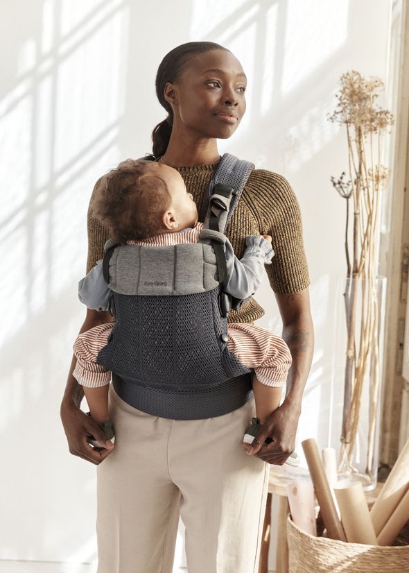 A woman stands near a sunlit window wearing a brown sweater, holding a baby in a carrier on her chest, with dried flowers and rolled papers nearby.