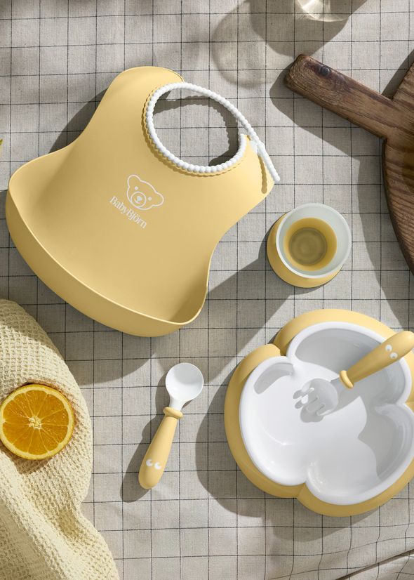 Yellow baby feeding set with a bib, divided bowl, cup, spoon, and a half-cut orange on a checkered tablecloth.