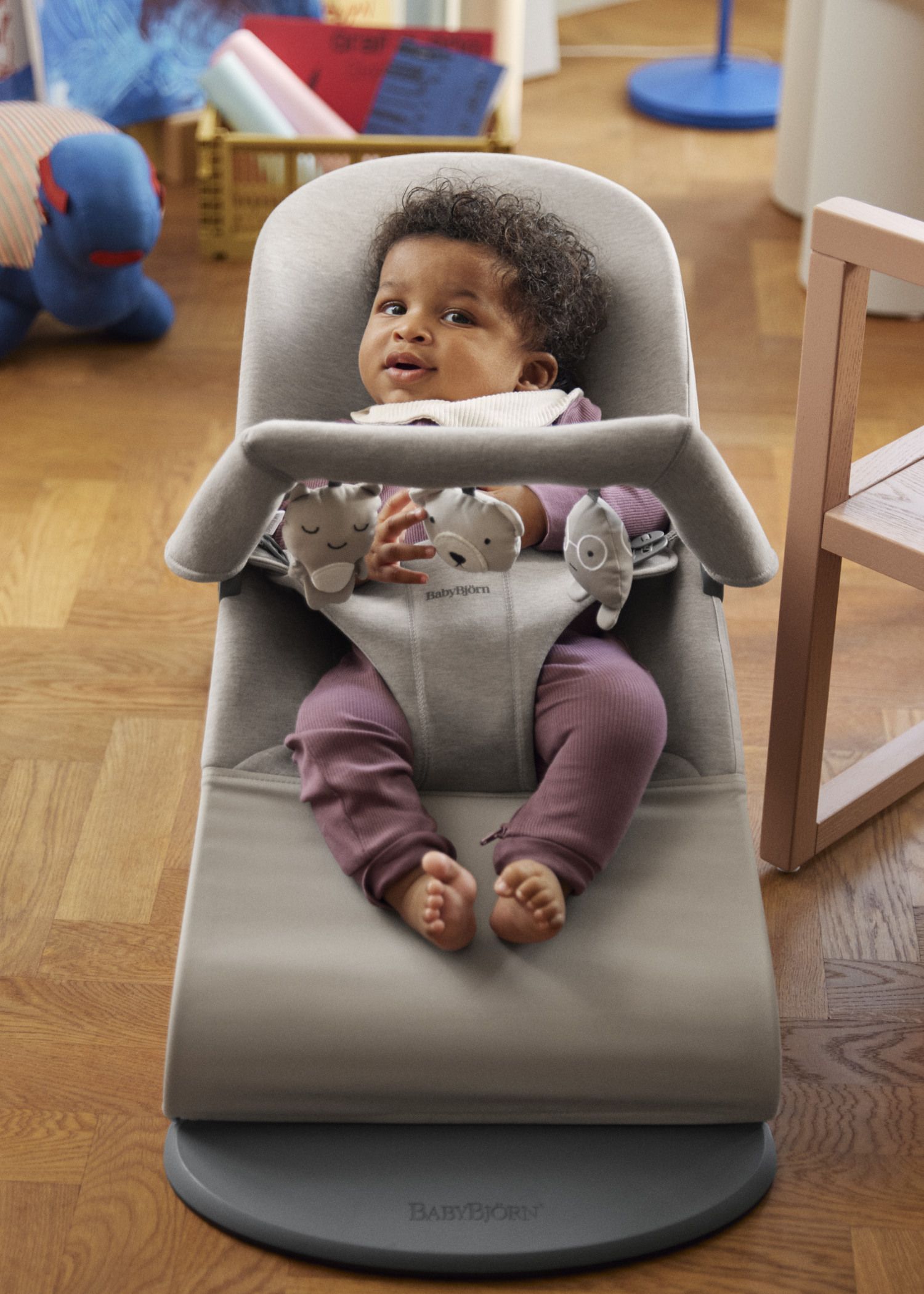 A baby in a purple outfit sits in a gray baby bouncer, surrounded by toys on a wooden floor.