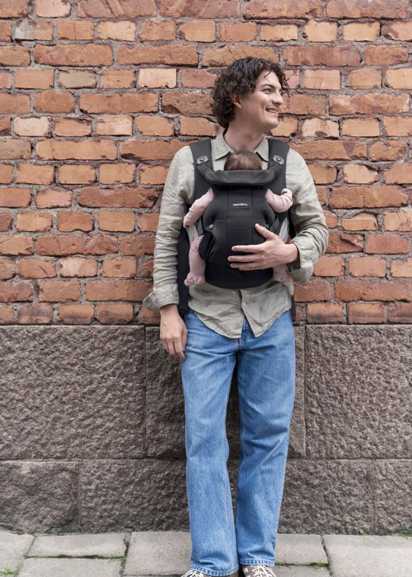A person smiling, standing against a brick wall, wearing a baby carrier with a baby inside, dressed in a light shirt and blue jeans.