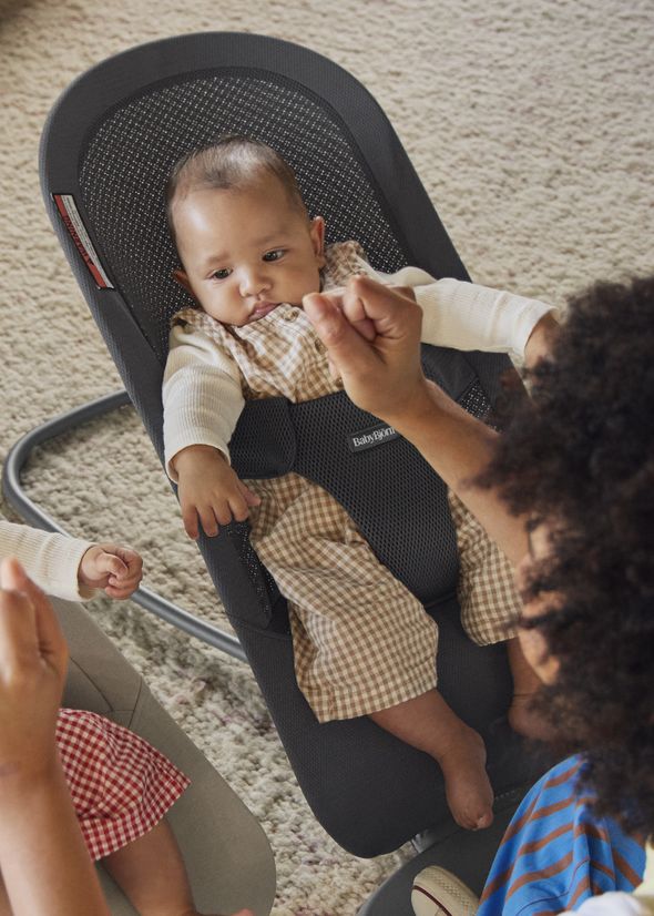 Baby in a bouncer wearing a checkered outfit, interacting with an adult on a cozy carpeted floor.