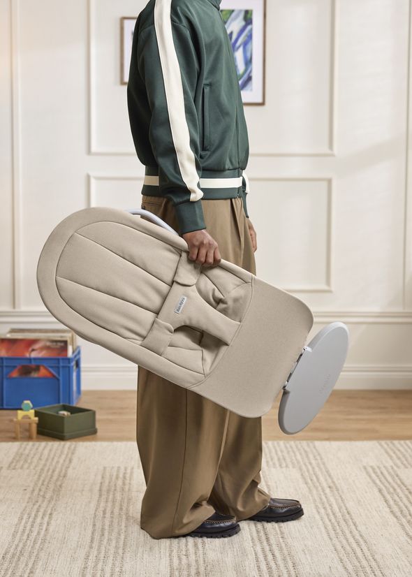 Person in a green and white jacket carrying a beige baby bouncer in a cozy room with toys on shelves.