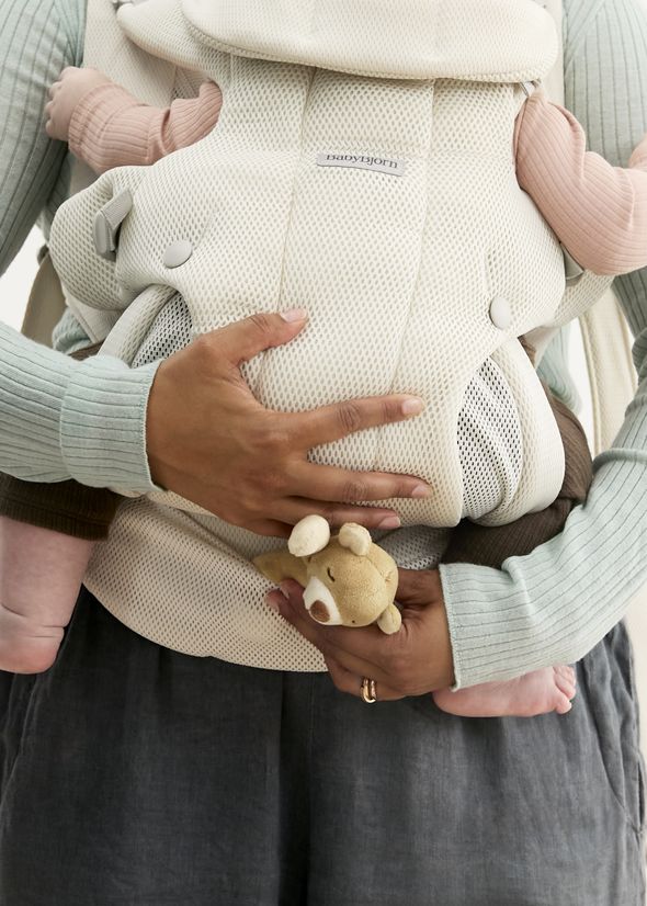 A person holds a baby in a white mesh Hold carrier from BabyBjörn, with a soft toy in hand. The baby wears pink and has tiny feet visible.