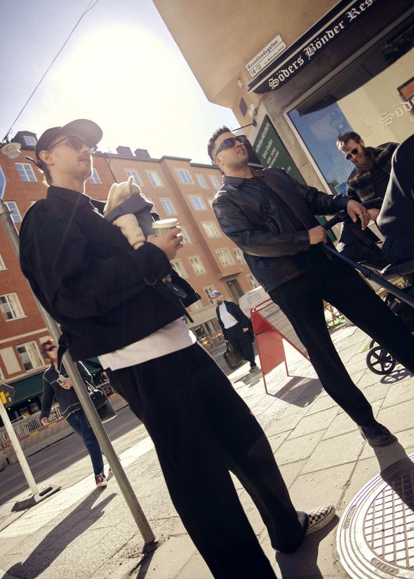 People standing on a sunny street corner, one holding a baby and a coffee, another with a stroller, near Söders Hjärta restaurant.
