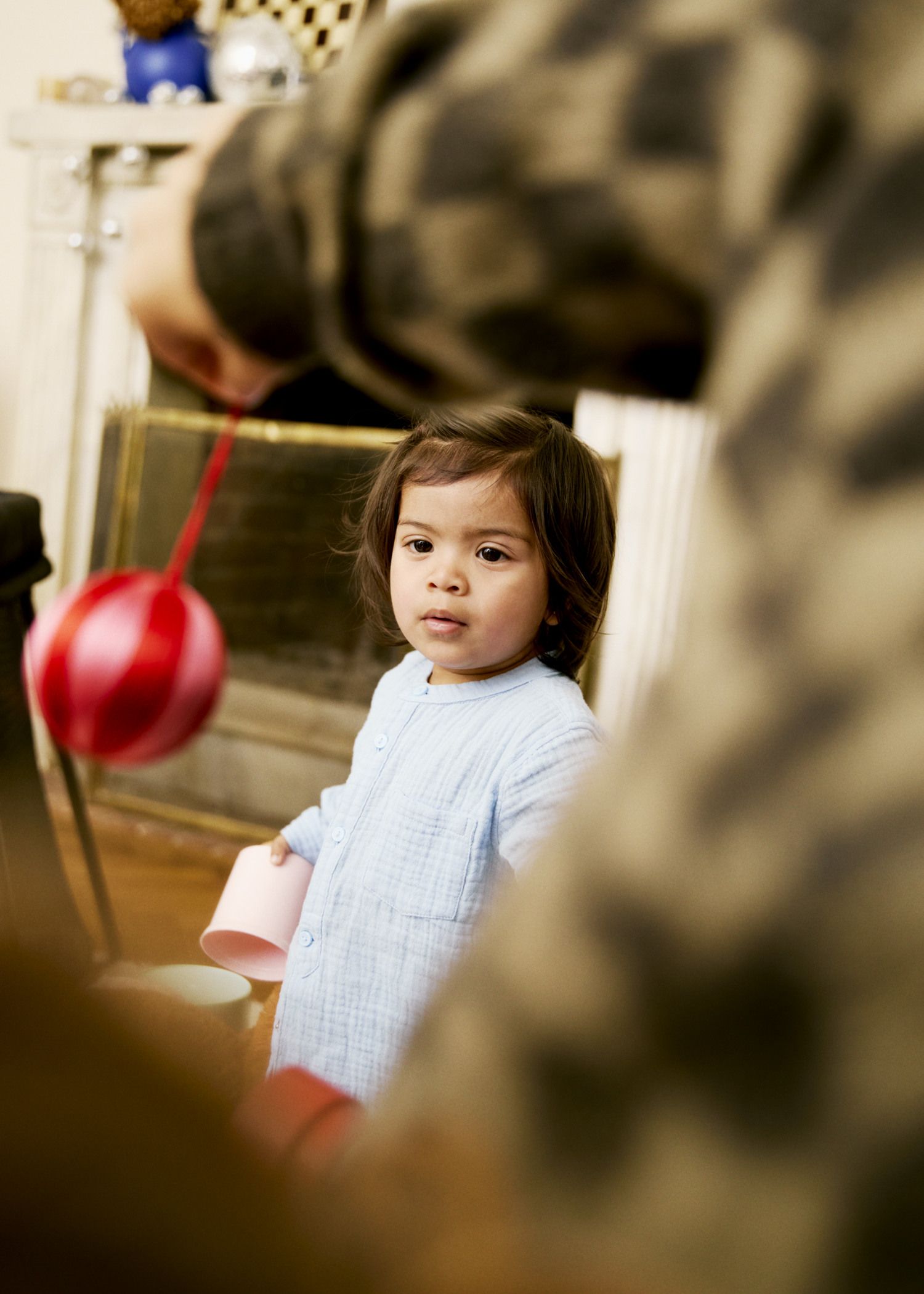 A child in a light blue outfit looks curiously at a red ornament held by an adult, standing in a room with a fireplace in the background.