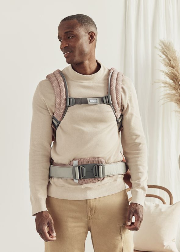 Man wearing a beige sweater and khaki pants models a baby carrier harness in a softly lit, modern interior setting.
