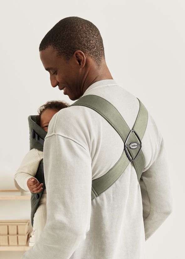 A person in a light sweater carries a baby in a green baby carrier with crossed straps, facing forward and smiling gently.