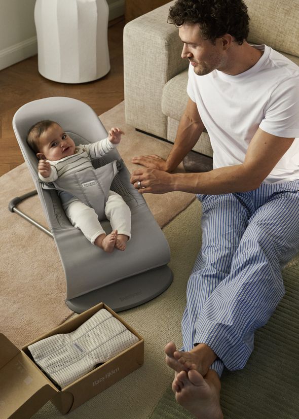 A man sits on the floor, smiling at a baby in a gray baby seat. Nearby is an open box with a blanket inside.