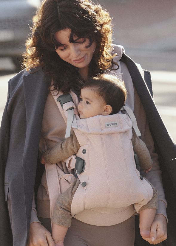 A woman smiles while carrying a baby in a pink baby carrier. She wears a coat draped over her shoulders.