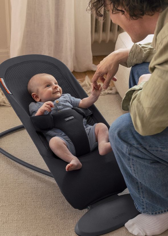 A smiling baby in a bouncer holds an adult's hand, showcasing a tender moment. The baby wears a blue outfit.