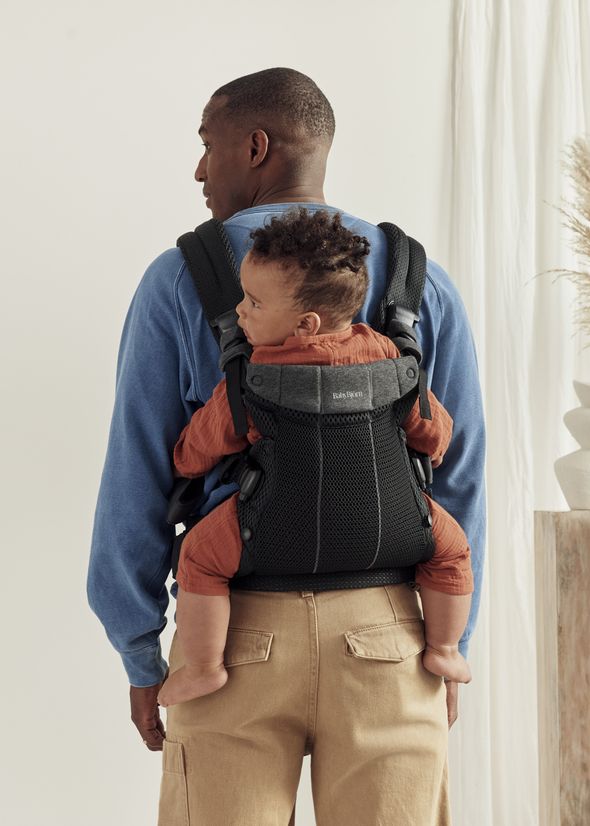 A person wearing a blue sweater carries a baby in a black baby carrier on their back, standing in a light-filled room with a plant.