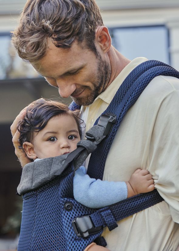 A man smiles down at a baby he is carrying in a blue baby carrier. The baby looks directly at the camera, dressed in a blue sweater.
