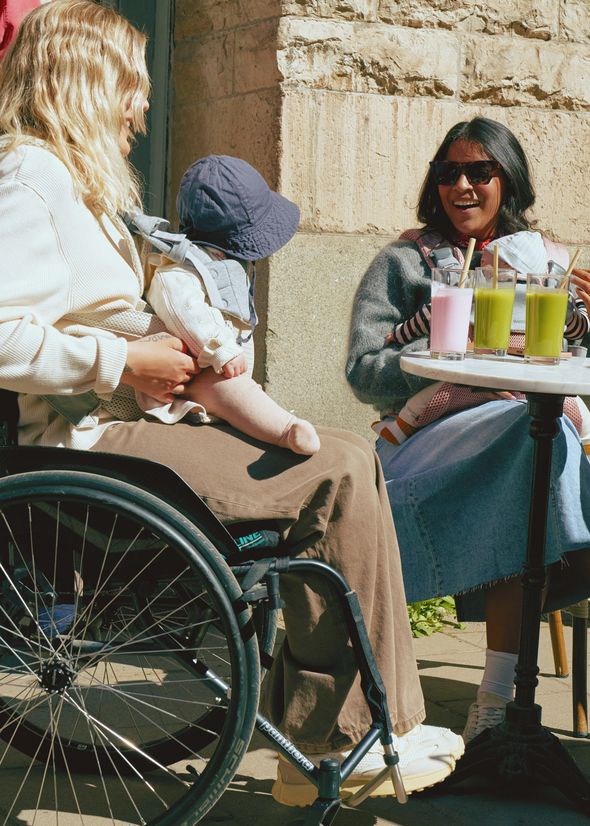 Two women sitting outdoors, one in a wheelchair holding a baby, the other at a table with drinks. Both are smiling and enjoying the sunny day.