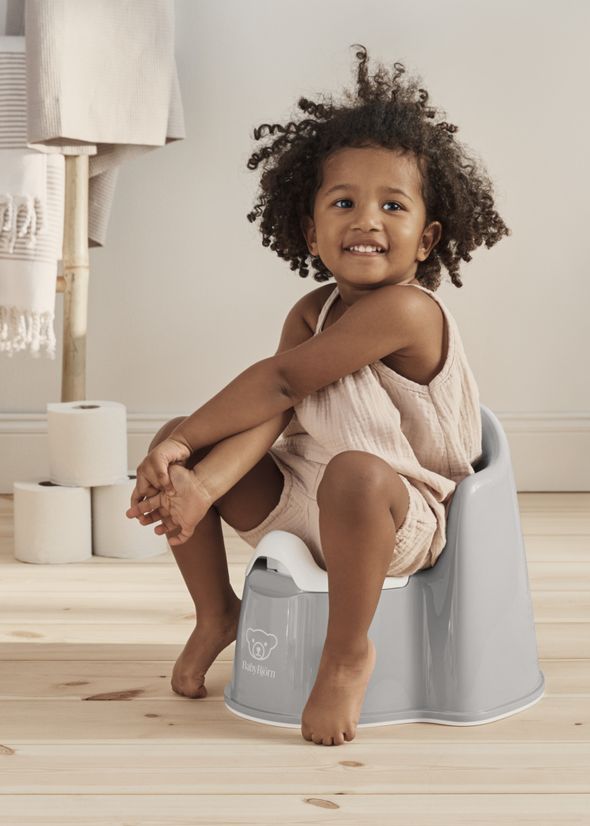 A child with curly hair sits on a gray potty chair, smiling. The room has wooden floors, rolled-up towels, and toilet paper in the background.