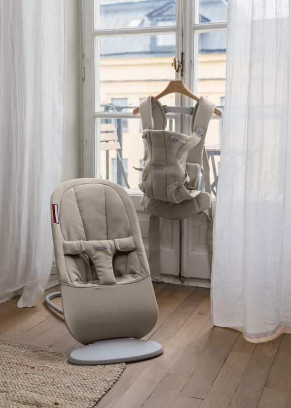 A beige baby bouncer and carrier are placed in a bright room by a window with sheer curtains, showcasing a cozy, minimalist design.