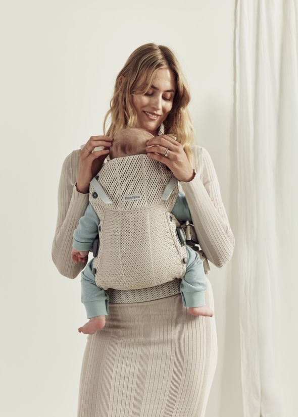 A woman smiles while holding a baby in a beige carrier. She wears a ribbed dress, and the baby is in a light blue outfit. A curtain is in the background.