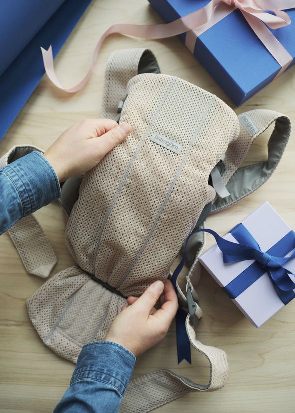 Person holding a beige baby carrier next to blue gift boxes with ribbons on a wooden surface.