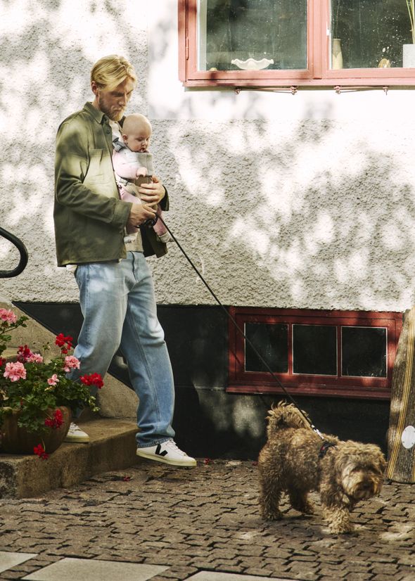 A person holds a baby while walking a small dog on a leash outside. The scene includes a building with a window and potted flowers.