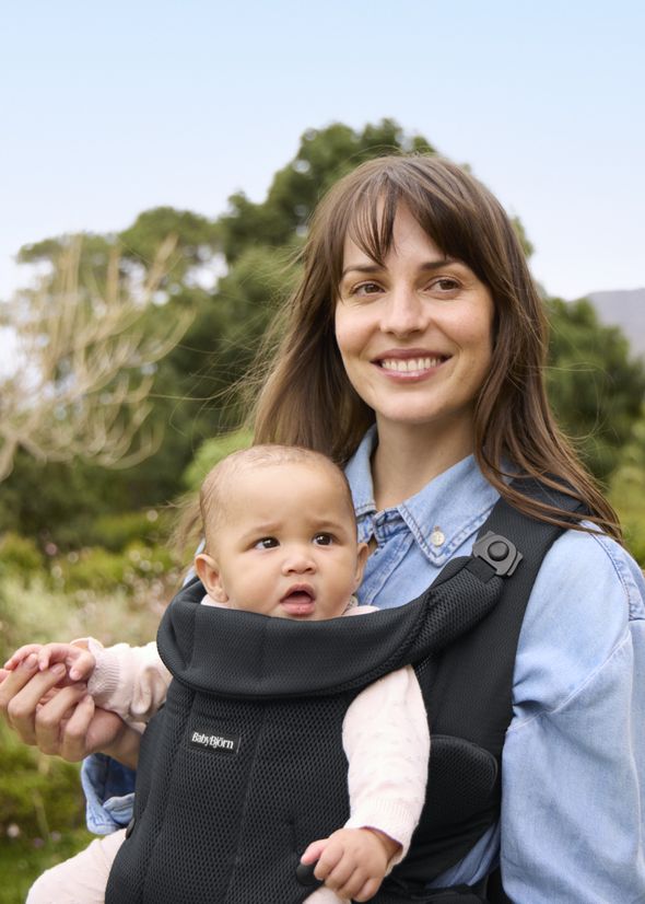 A woman smiles while holding a baby in a carrier, standing outdoors in a park with greenery and trees in the background.