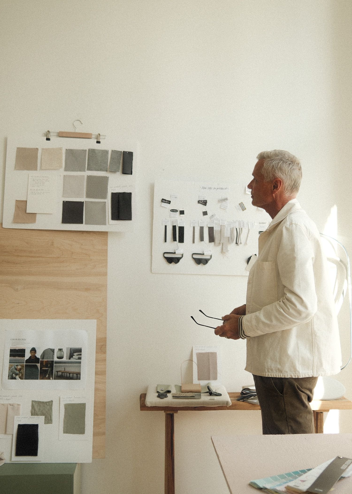 A person standing in a studio, looking at fabric swatches and design boards on the wall, holding glasses in hand, with sunlight streaming in.