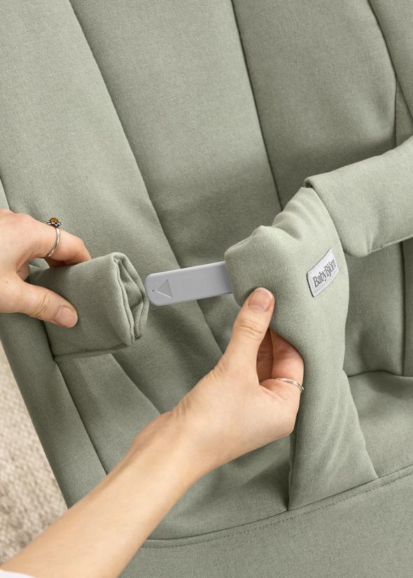 Hands adjusting a light green baby bouncer with a BabyBjörn label, showcasing a strap and padded cover.