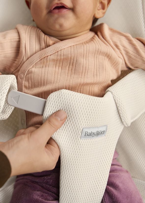 A baby in a peach shirt is secured in a BabyBjörn bouncer with a hand adjusting the white mesh harness.