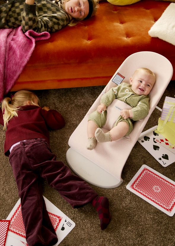 A baby in a bouncer smiles, surrounded by playing cards. Two children lie on the floor nearby, one in a red sweater and pants.