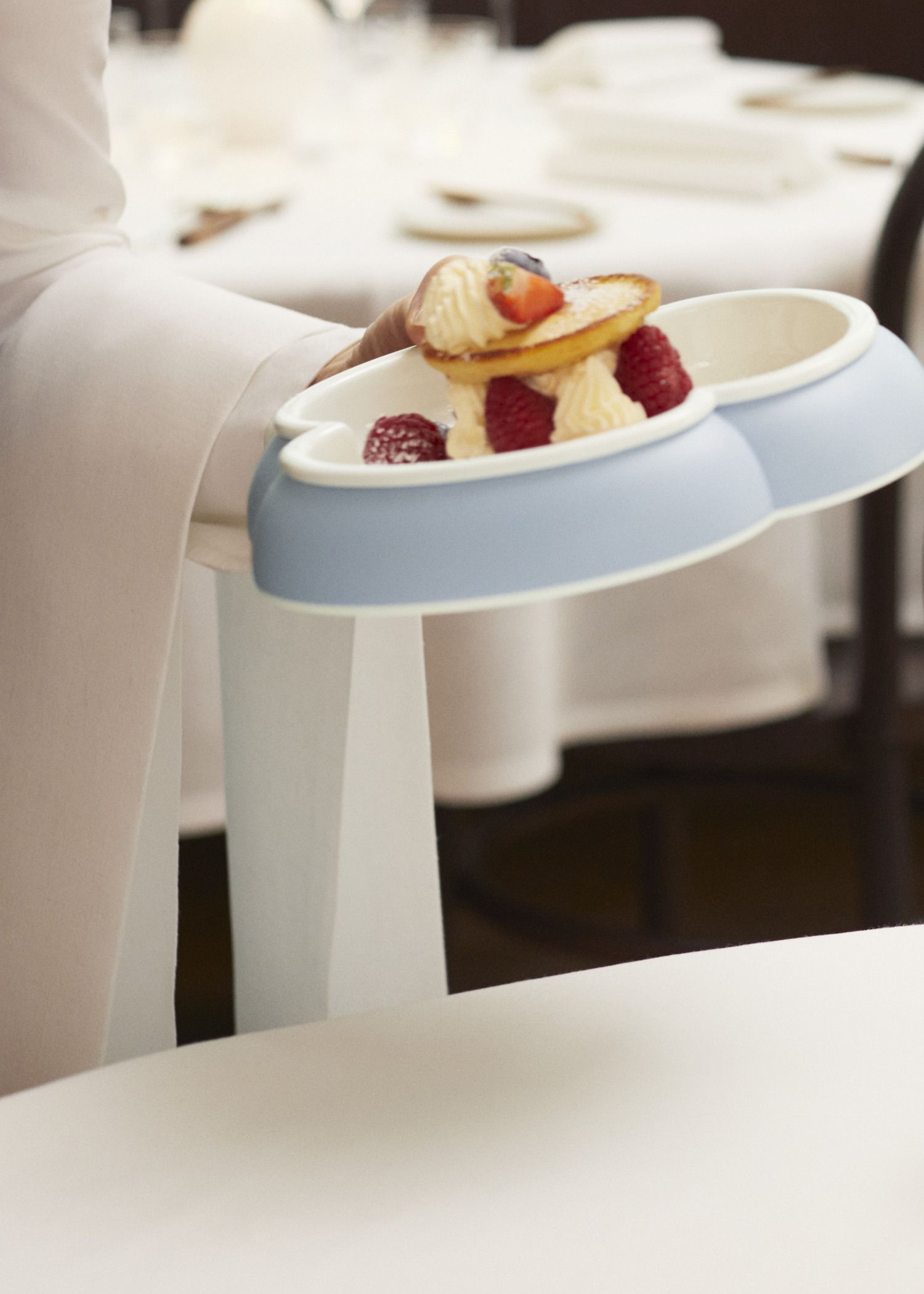 A person holds a blue tray with desserts topped with raspberries and strawberries, with a blurred dining setting in the background.