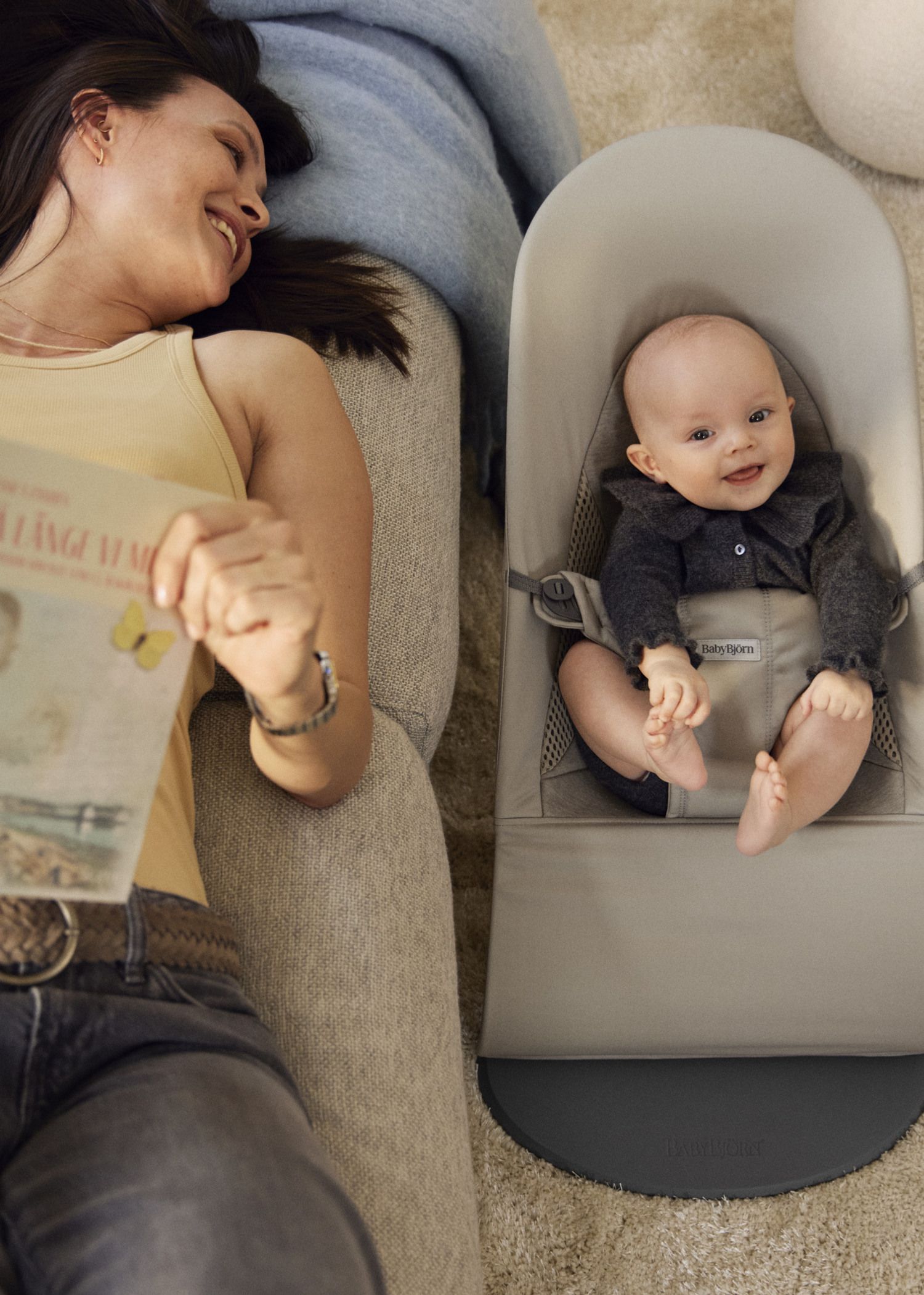 A woman relaxes on a couch holding a magazine, smiling at a baby in a gray bouncer. The baby is smiling back, wearing a dark outfit.