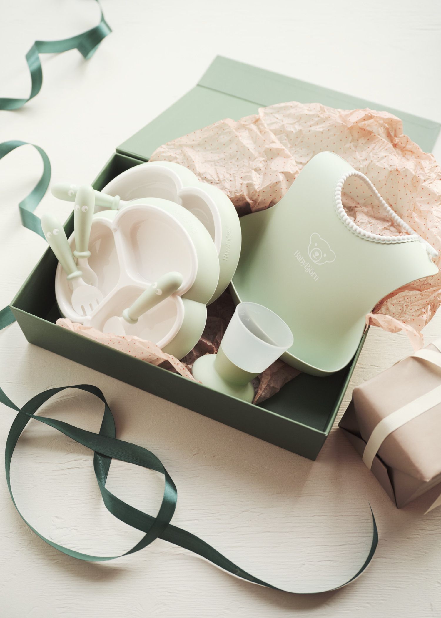 A gift box containing a green baby bib, divided plate, utensils, and cup, surrounded by pink tissue paper and ribbon on a light surface.