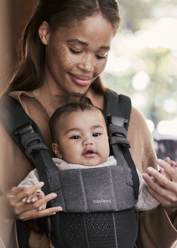 A woman smiling while holding a baby in a gray carrier. The baby is looking forward, and both appear content.