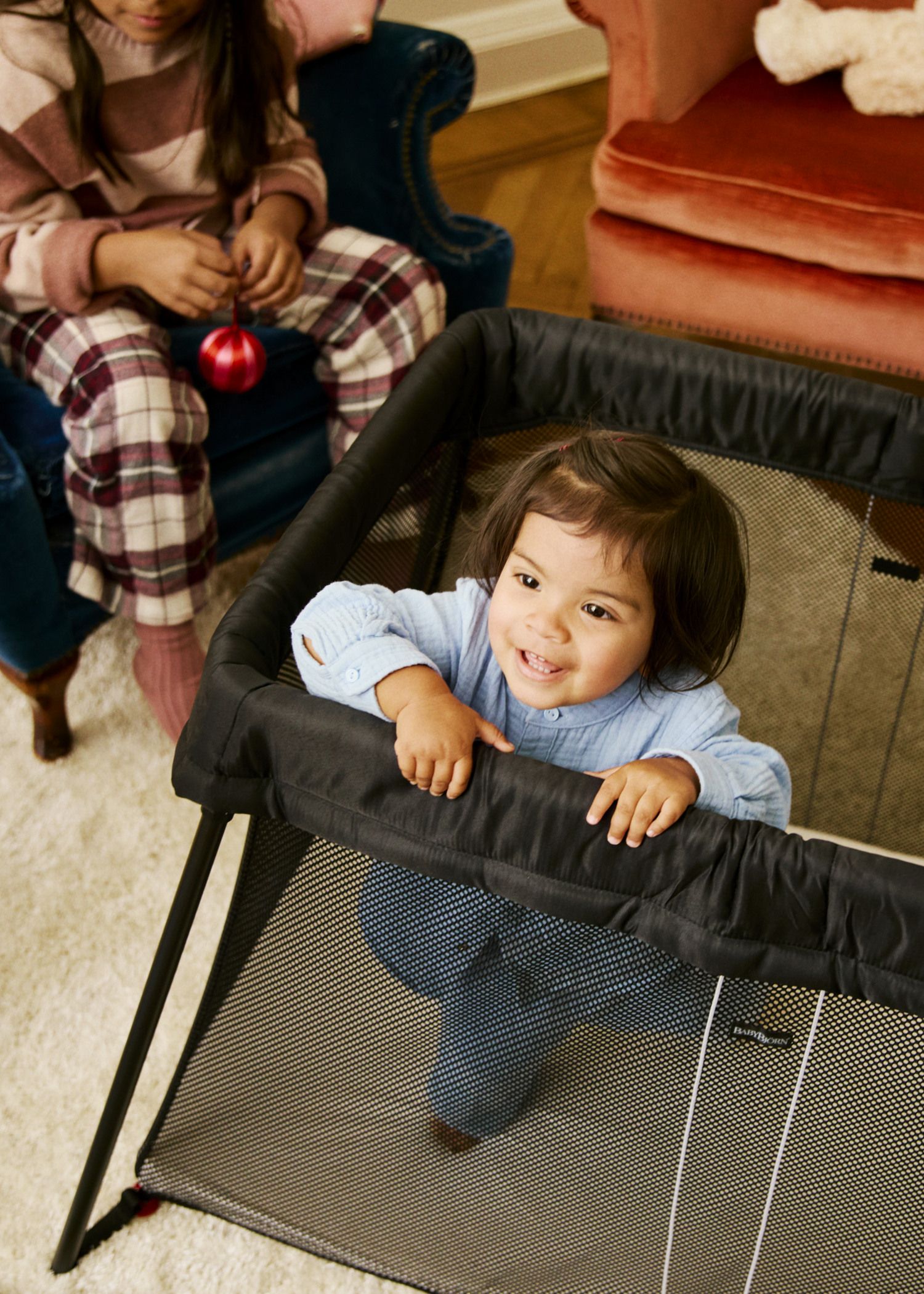 A smiling toddler in a blue outfit stands in a playpen. A child in plaid pajamas sits nearby holding a red ornament.