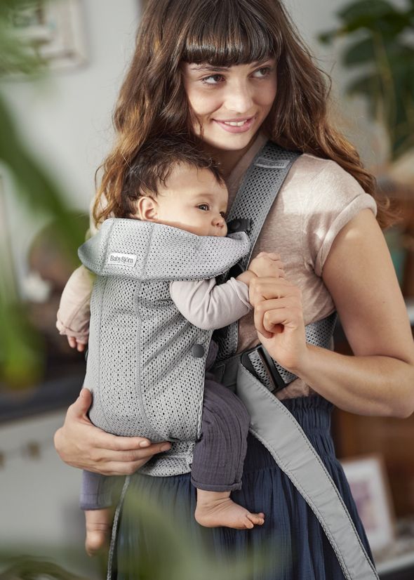 A woman smiles while holding a baby in a gray BabyBjörn carrier indoors. The baby rests comfortably.