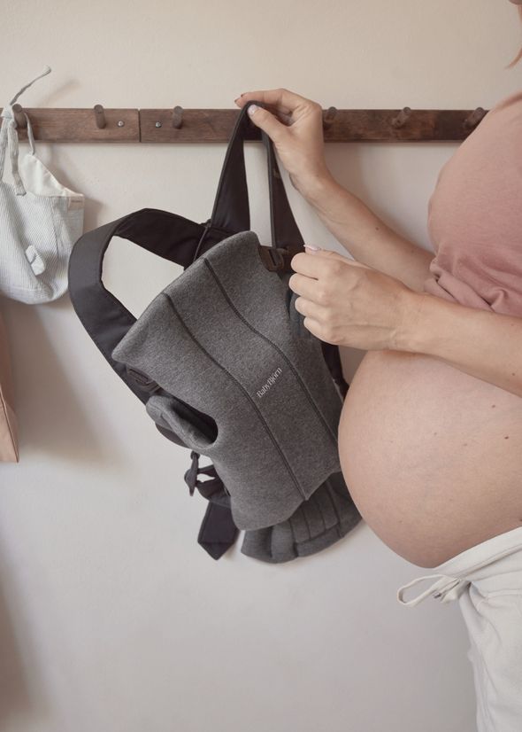 Pregnant woman holding a grey baby carrier, hanging it on a wooden hook, with her belly visible.