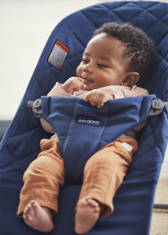 A baby in a blue bouncer chair, wearing a light pink shirt and brown pants, looking content and relaxed.