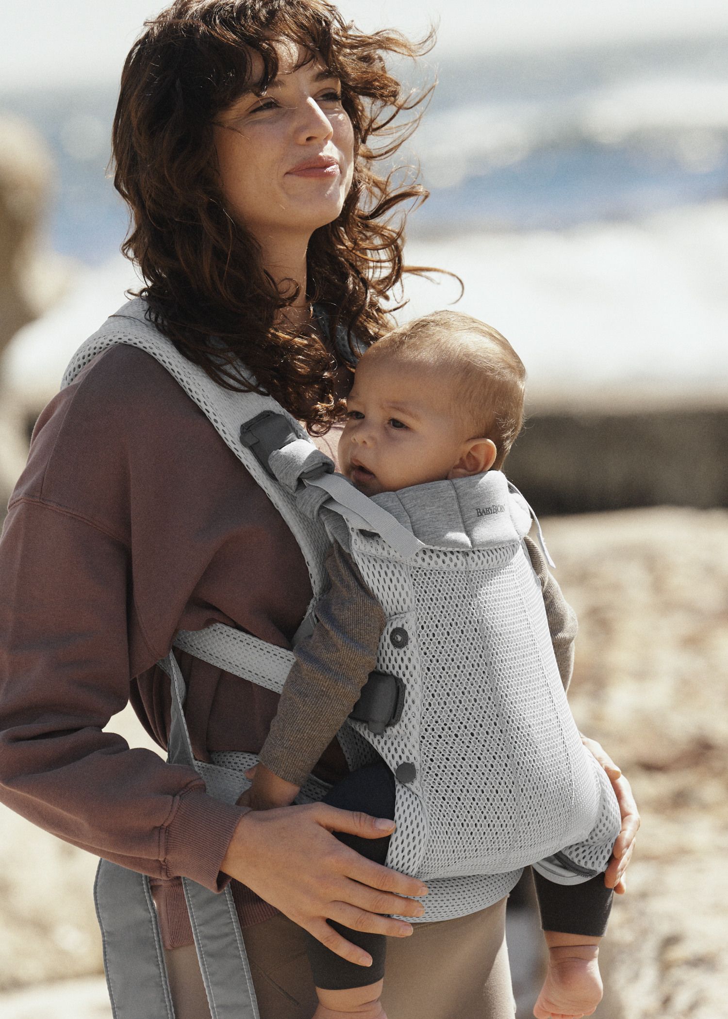 Woman outdoors carrying a baby in a gray baby carrier. Both are looking into the distance, with the ocean in the background.