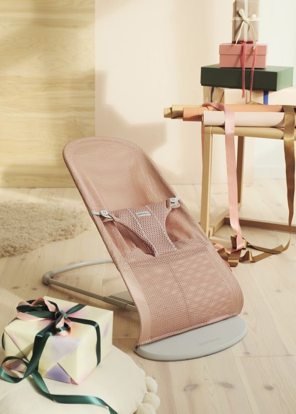 A pink baby bouncer on a wooden floor, next to a stack of colorful, ribbon-tied gift boxes on a chair.