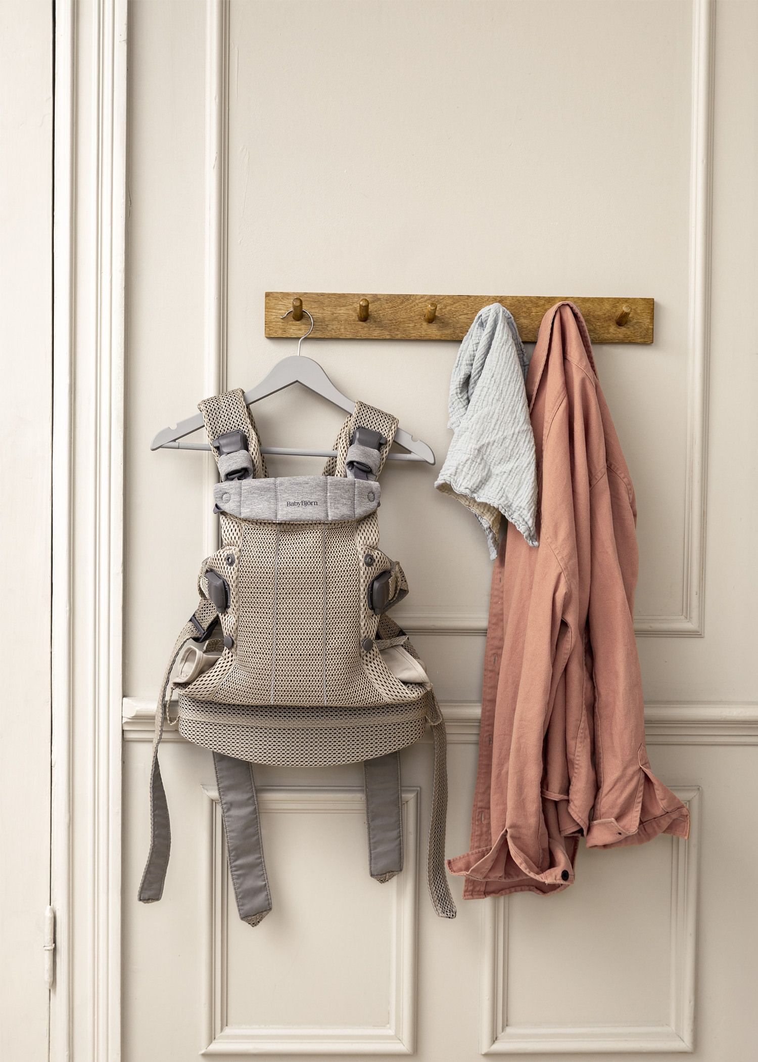 A beige baby carrier hangs on a wooden rack, alongside a light blue cloth and a pink shirt, against a cream-colored door.