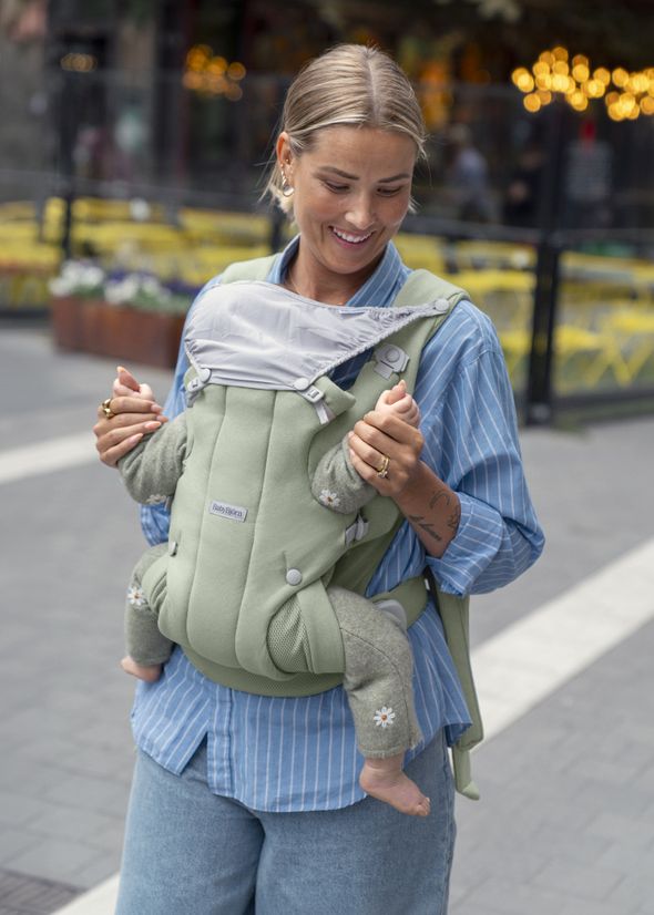 Woman smiling, wearing a blue-striped shirt, carrying a baby in a green carrier on a city street with a blurred outdoor café in the background.