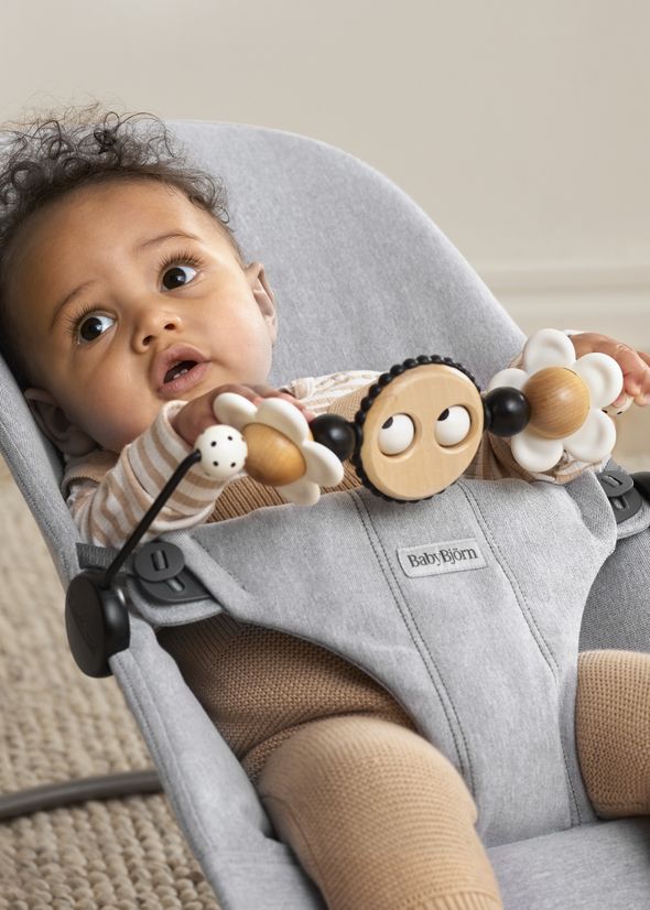 Baby in a grey bouncer seat, holding onto a toy bar with wooden and plastic elements, wearing a striped top and brown pants.
