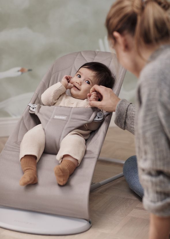 A baby in a light-colored onesie sits in a gray bouncer, smiling and holding an adult's hand, with a soft green background.