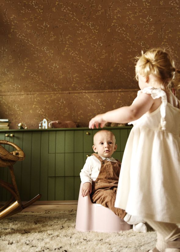 A baby sits on a pink potty, wearing brown overalls, while a toddler in a white dress stands nearby on a patterned rug in a cozy room.