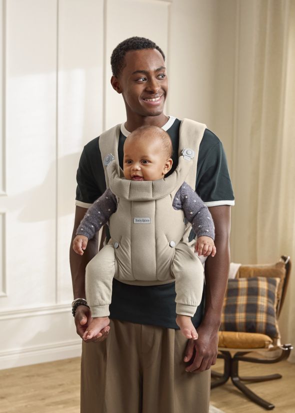 A smiling man stands in a room wearing a black shirt, holding a baby in a beige carrier. The baby is wearing a dark sweater.