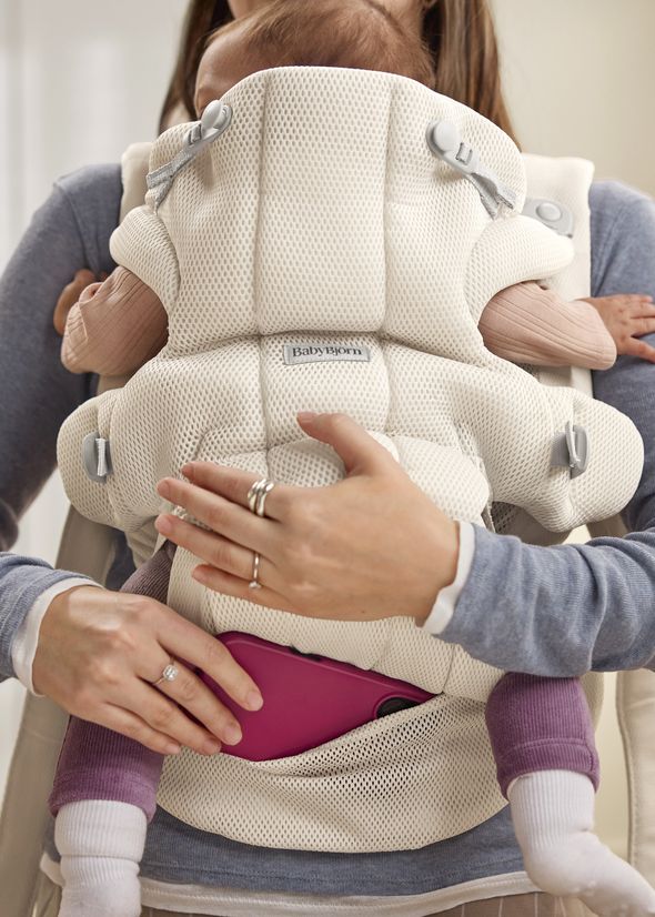 A baby is comfortably carried in a white mesh baby carrier, with an adult's hand adjusting the carrier and supporting the baby, who wears purple pants.