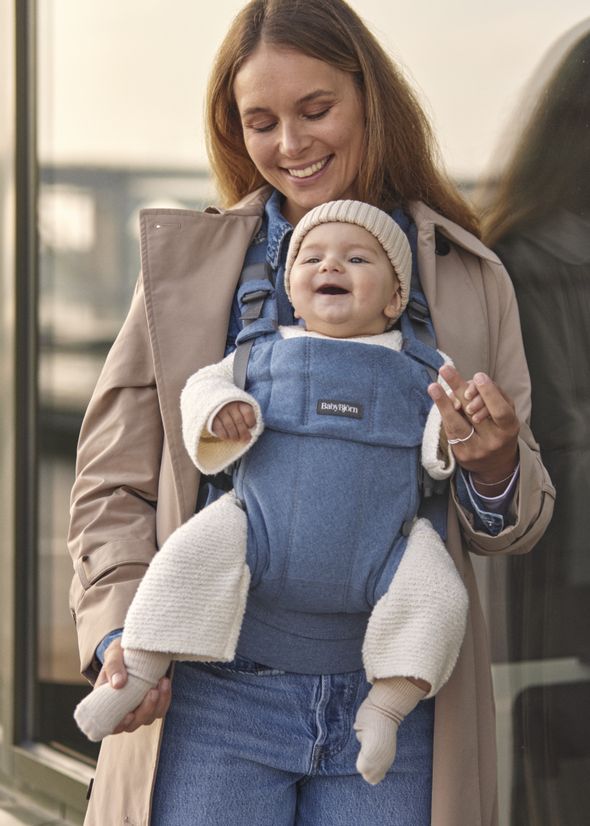 Smiling woman in a beige coat carries a happy baby in a blue carrier. The baby wears a knitted hat and booties.