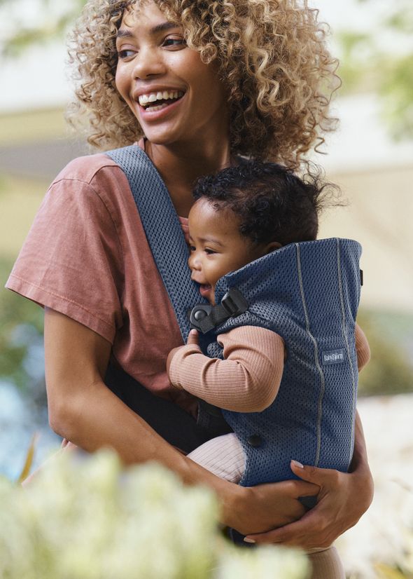 Smiling woman carrying a baby in a blue baby carrier outdoors. The baby is wearing a brown outfit.